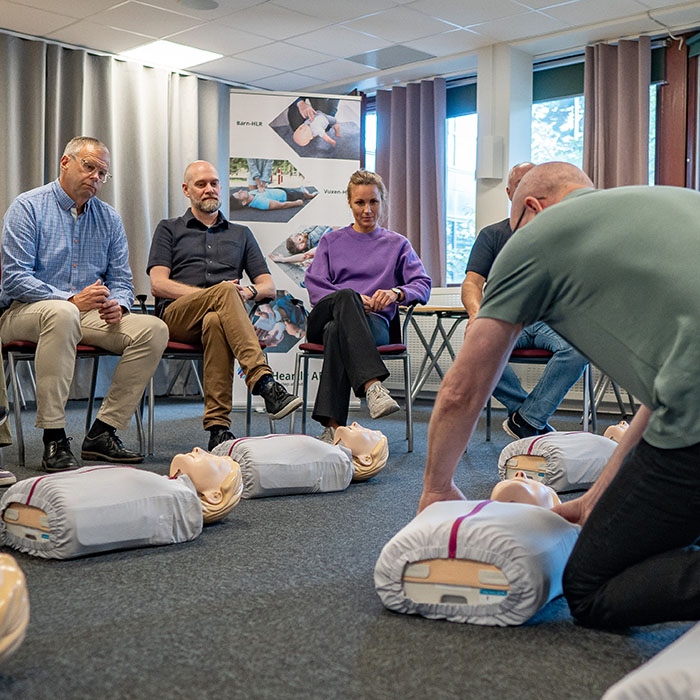 Manlig instruktör demonstrerar hur man utför HLR på en docka där elever sitter i en ring och följer med.